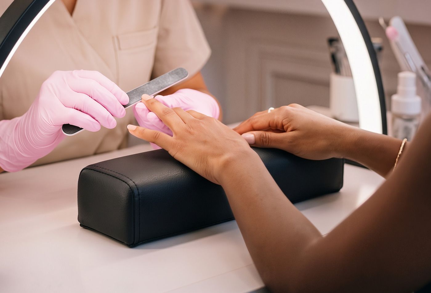 A client's hand rests on the black nail arm rest while a technician in pink gloves files a nail.