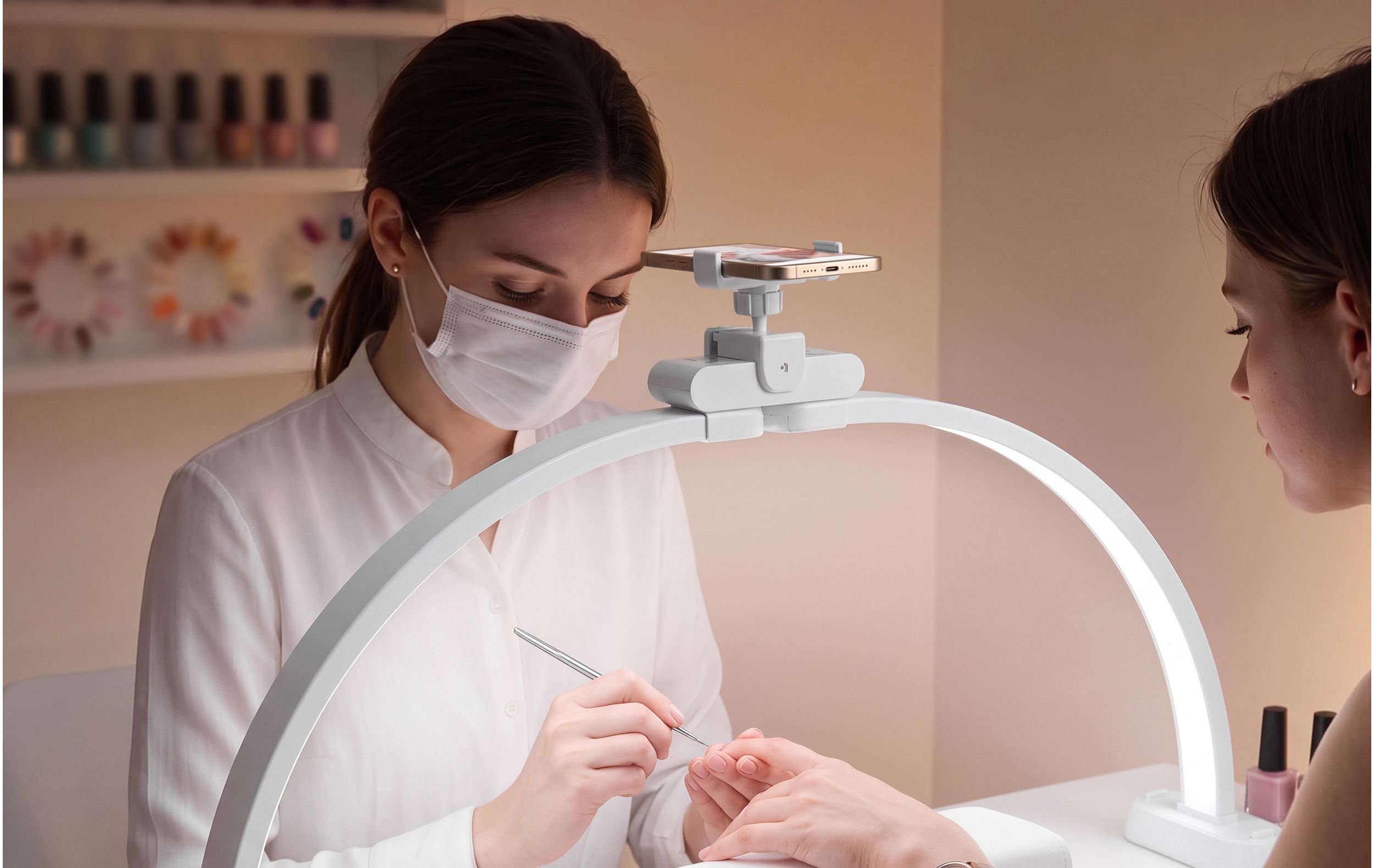 A nail technician works on a client's nails under the white Half-Moon Light, with a phone clipped into the holder above the control module.
